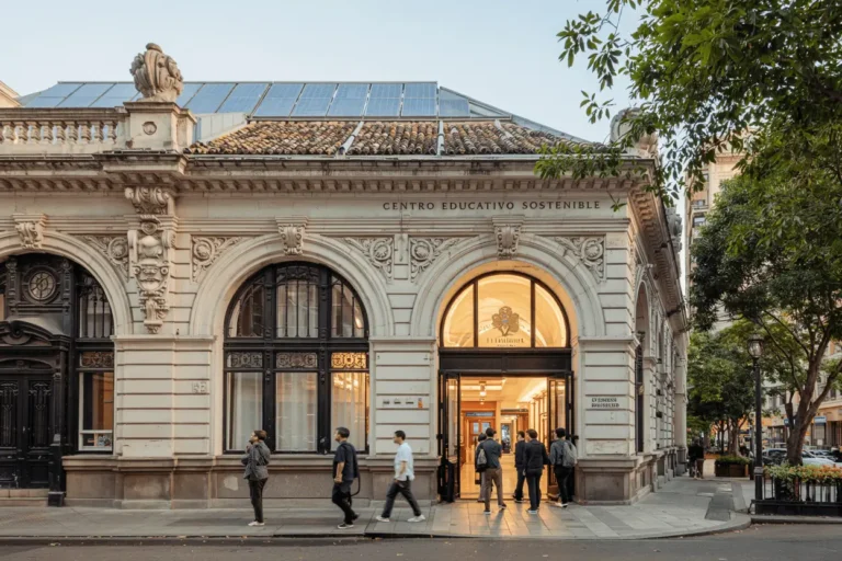 Historic Argentine building with neoclassical architecture transformed into a sustainable educational center, featuring solar panels on the roof while preserving ornate facade details. Students entering the illuminated "Centro Educativo Sostenible" demonstrate successful energy retrofit that balances cultural heritage preservation with modern sustainability practices.